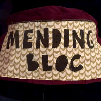 small apron with reverse applique saying "mending bloc" in a patterned white/green fabric over a solid burgundy fabric